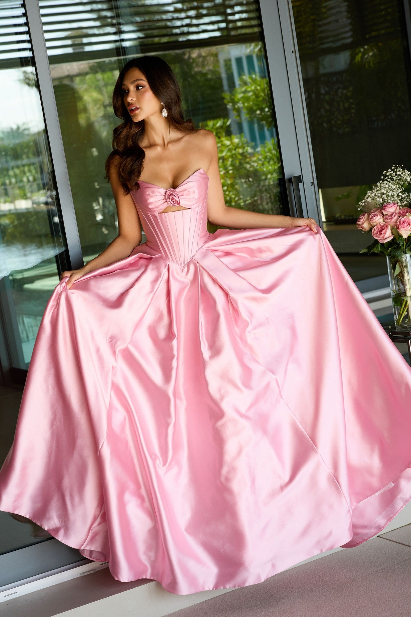 A woman indoors holds the sides of her pink Strapless Satin Prom Ball Gown (Ava Presley 47223), with a bouquet of pink roses on a table beside her.